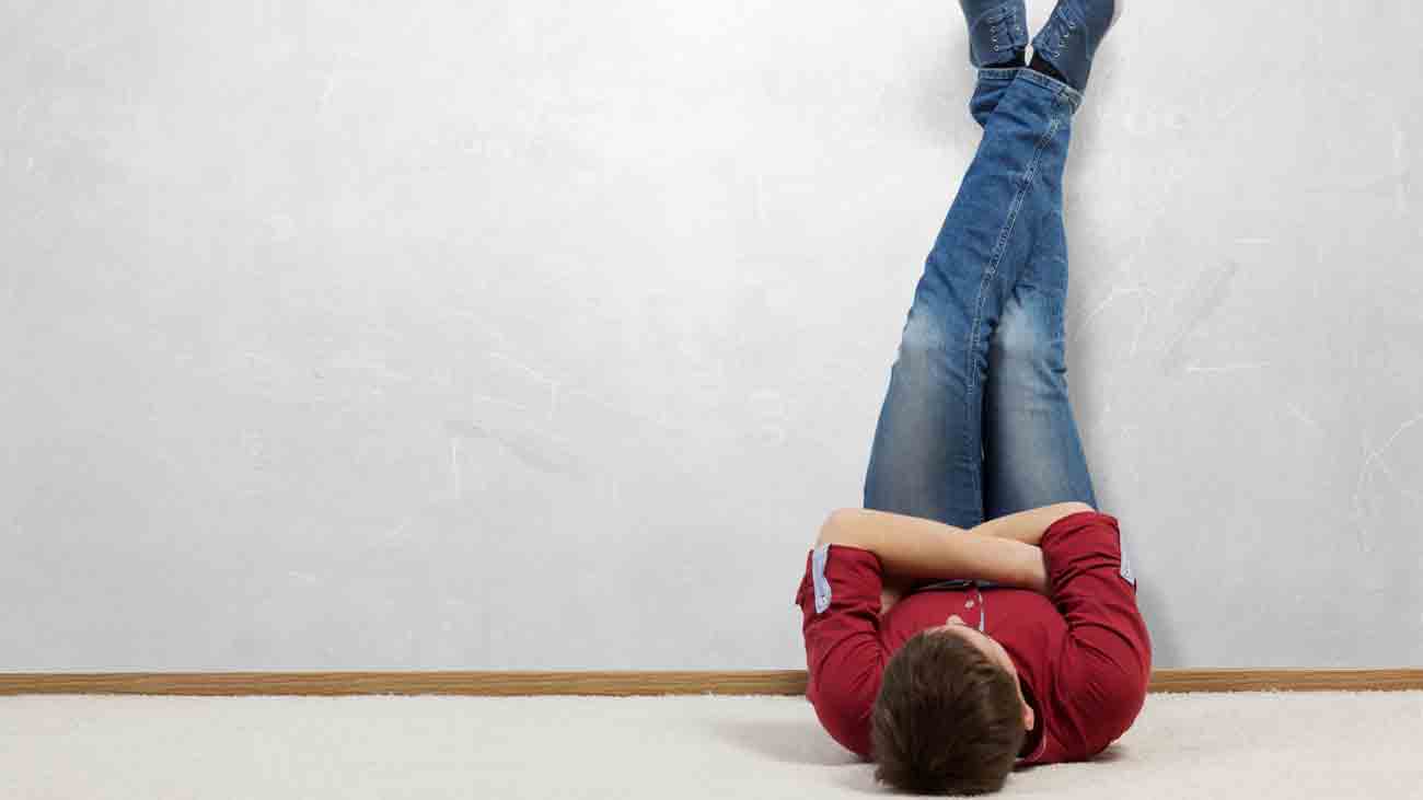 A young man in blue pants and a red shirt is doing yoga with his legs up against the wall.