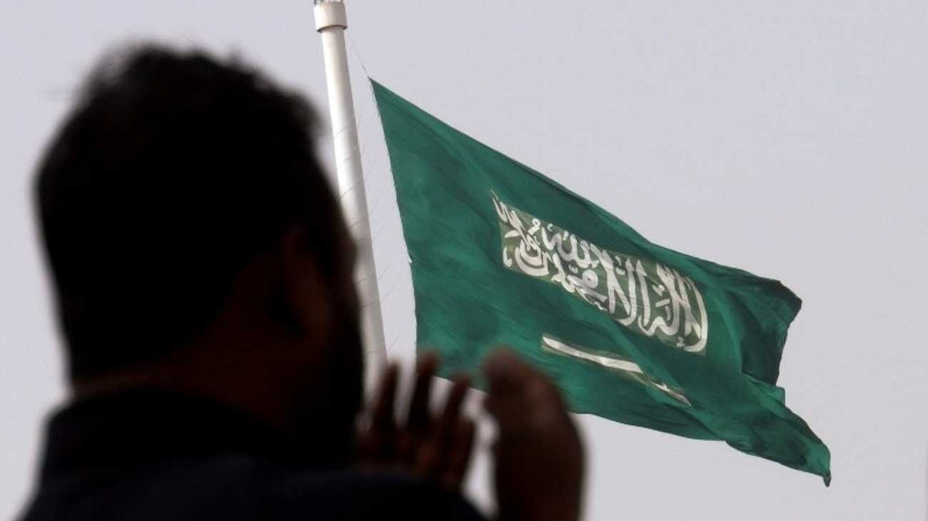 ایک شخص سعودی عرب کے جھنڈے کے سامنے کھڑا ہو کر دعا مانگ رہا ہے